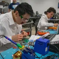 Individuals work on electronic projects in a classroom setting using tools like soldering irons and multimeters.