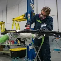A person uses a power sander on a car part in an automotive repair shop with various tools and equipment around.