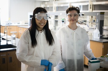 Two STEM students wearing white lab coats and safety goggles in a lab