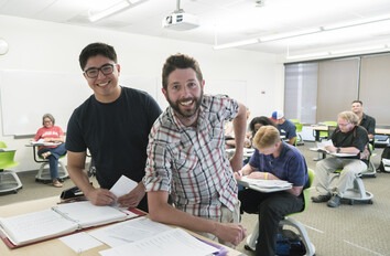 Two people standing at a desk in the front of a classroom smiling at camera