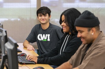 Three students sitting in a row in a computer classroom and one is looking at the camera