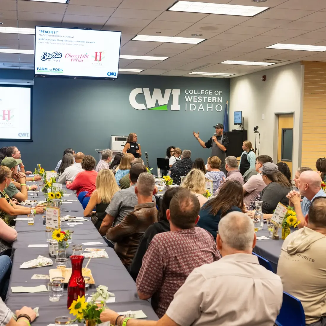 Rows of people sit in front of tables, while watching speaker