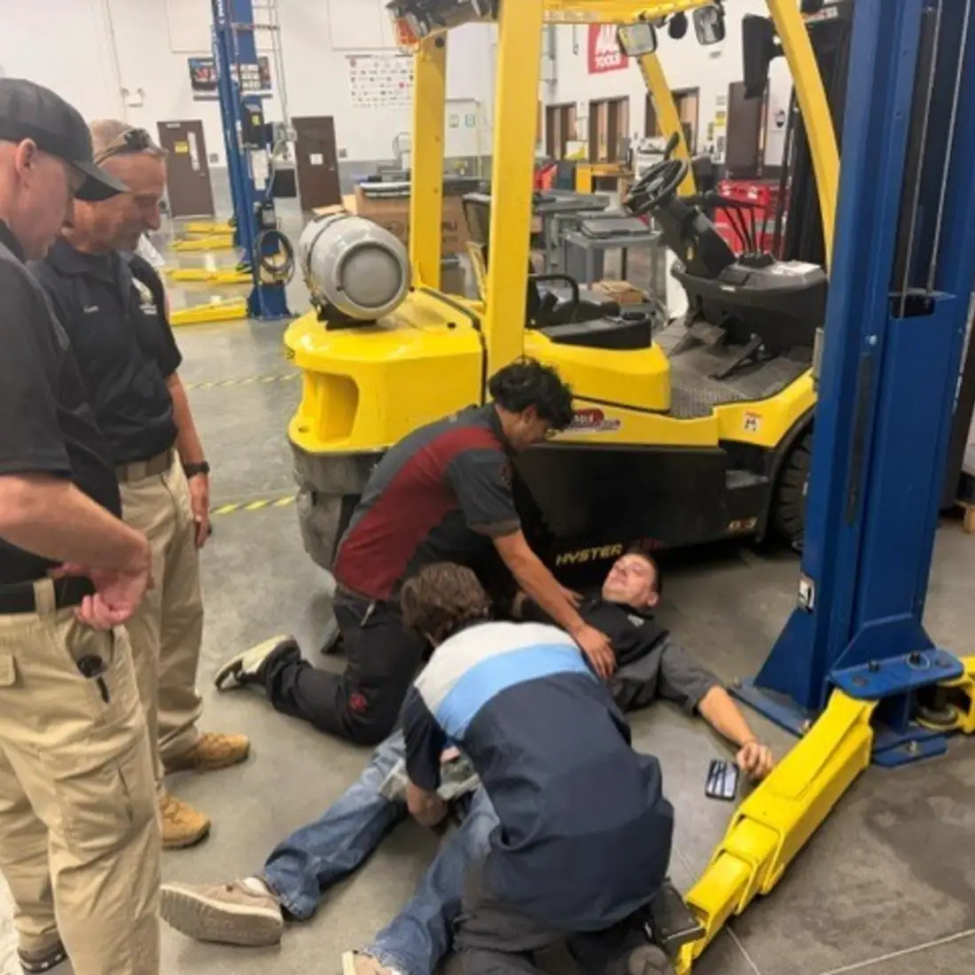 Workers assist a person lying beside a forklift as colleagues observe