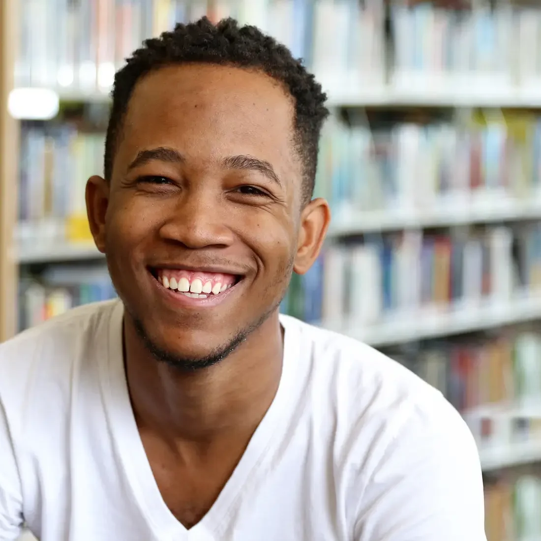 Happy student from Jamaica wearing a white t-shirt sitting in front of shelf of books