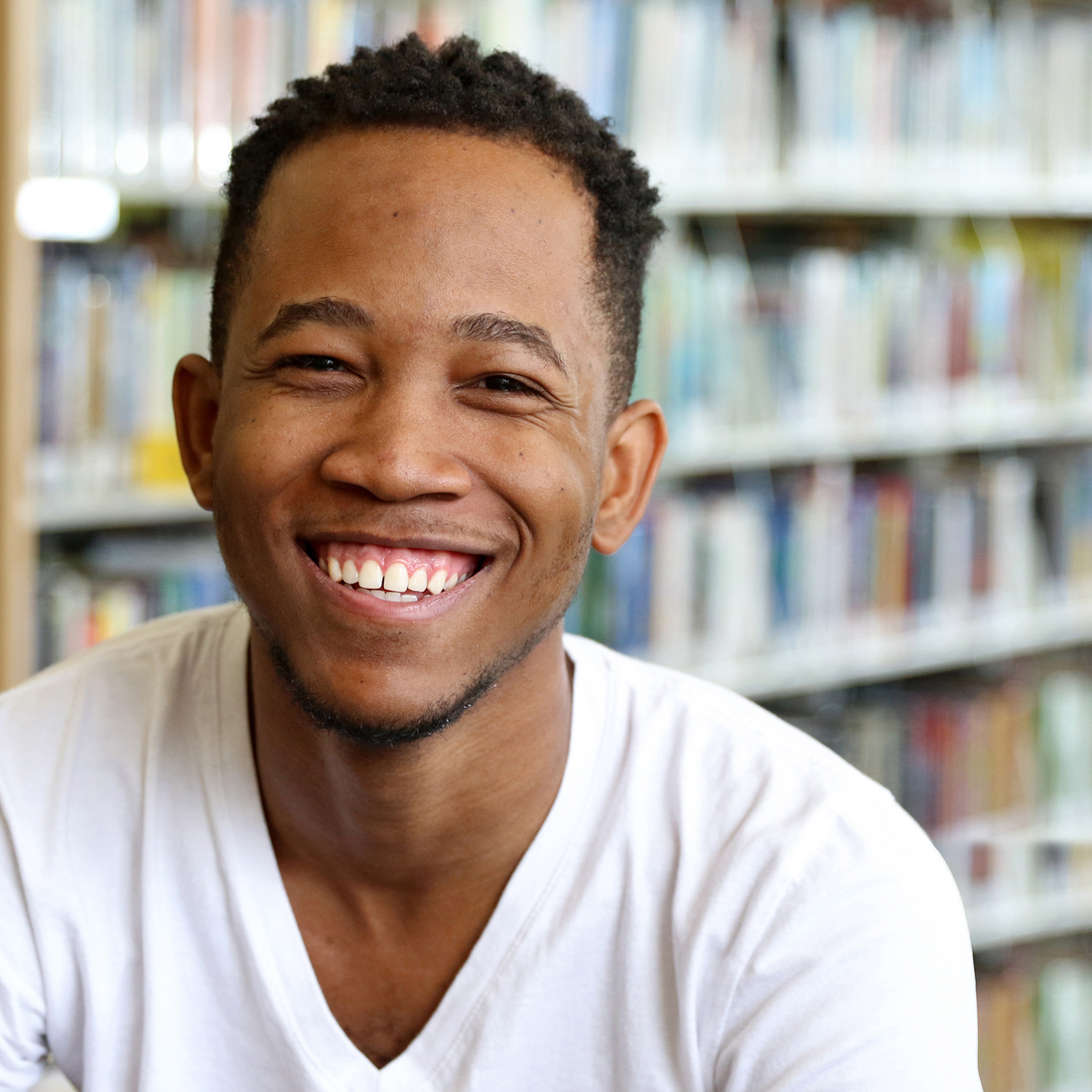 Happy student from Jamaica wearing a white t-shirt sitting in front of shelf of books