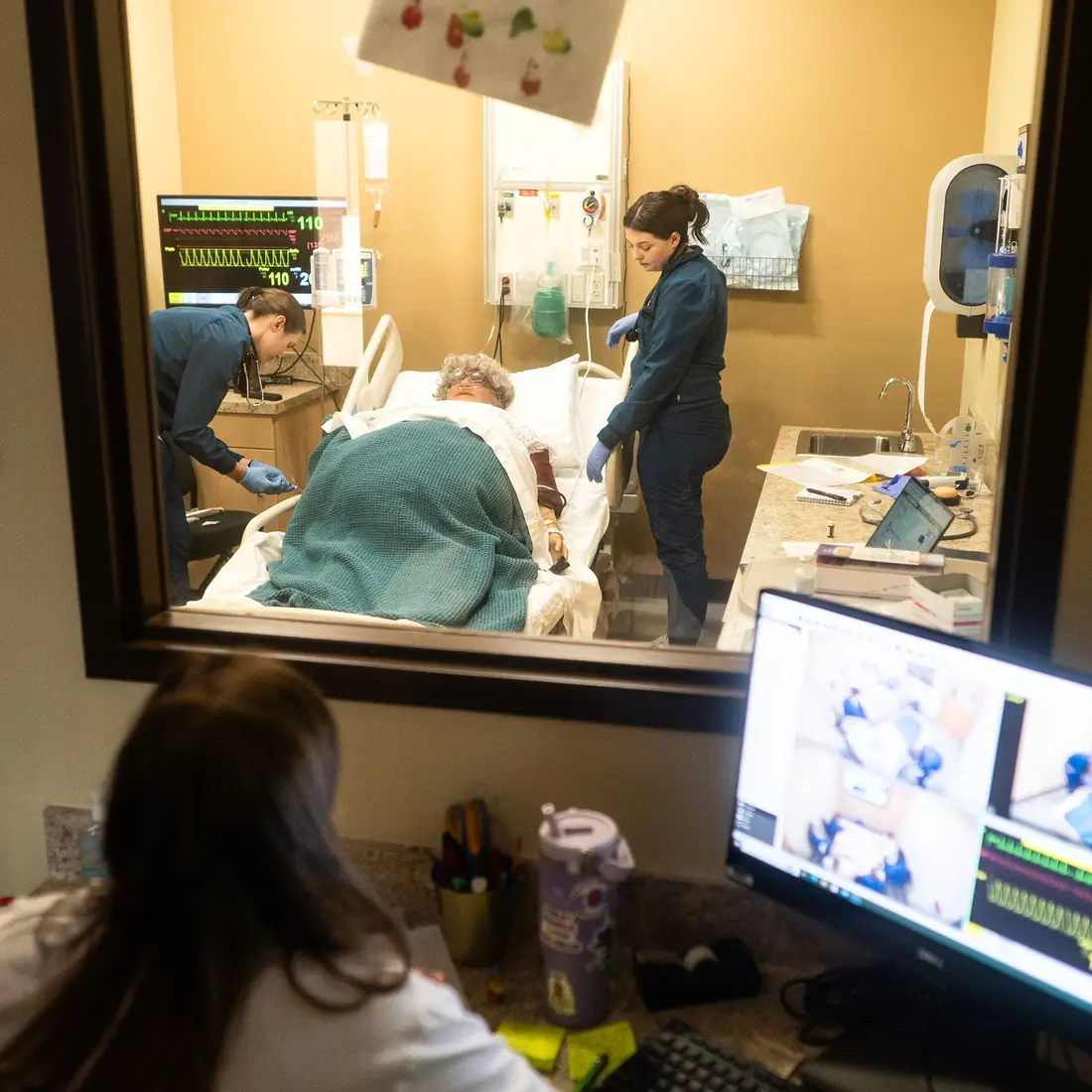 Two nursing students learn in a simulated hospital classroom as a teacher observes through a pane of glass.