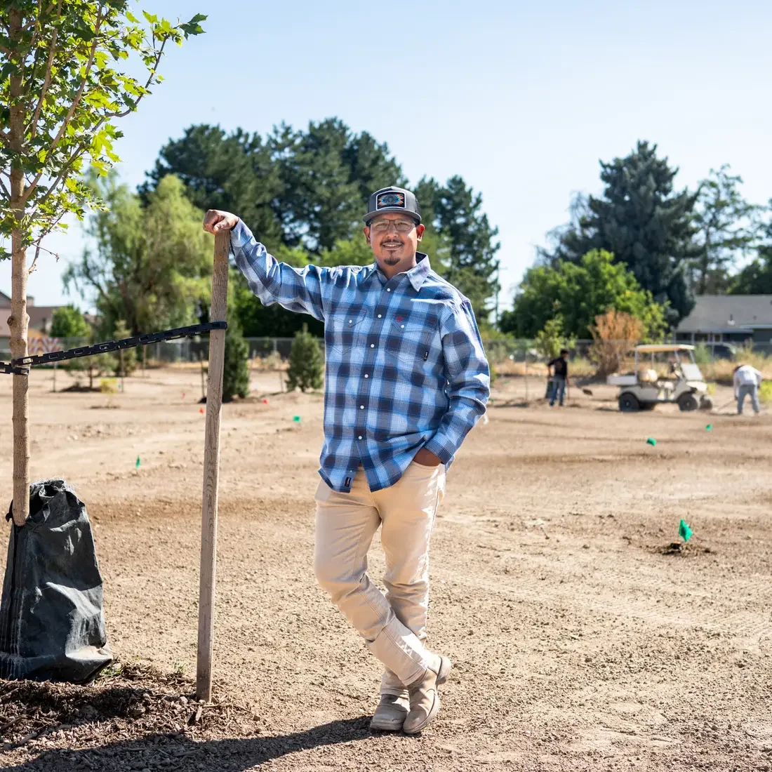 A man stands in a field with newly planted trees and employees working behind him.