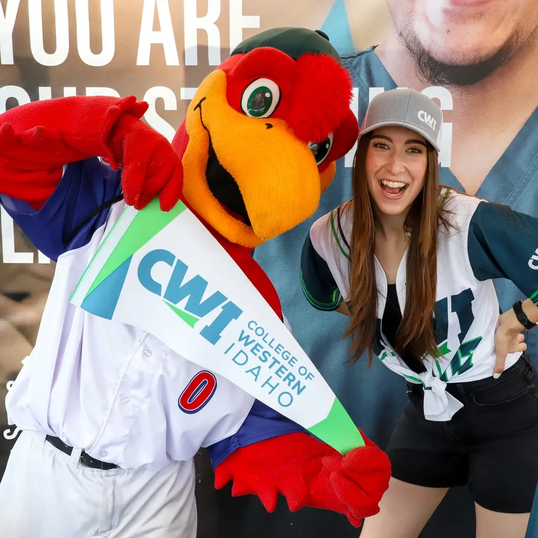 A woman wearing a College of Western Idaho baseball jersey smiles as she poses next to the Idaho Hawks mascot.