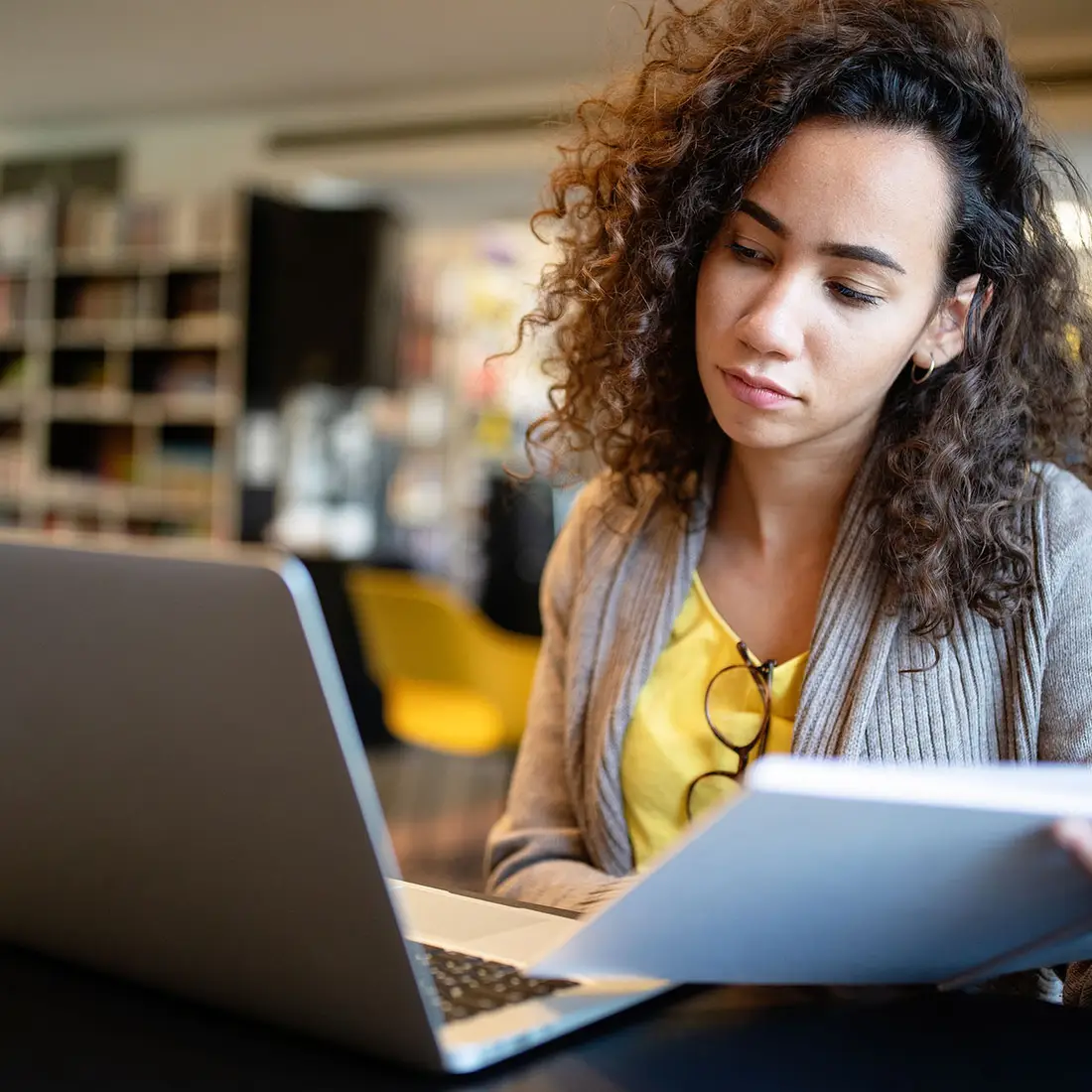 A college student sits in a library in front of a laptop while holding a notebook.