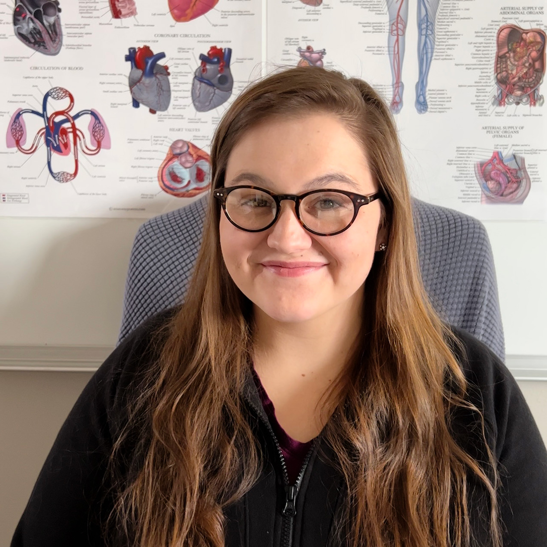A portrait of Alex Pless sitting in front of an Anatomy and Physiology poster.