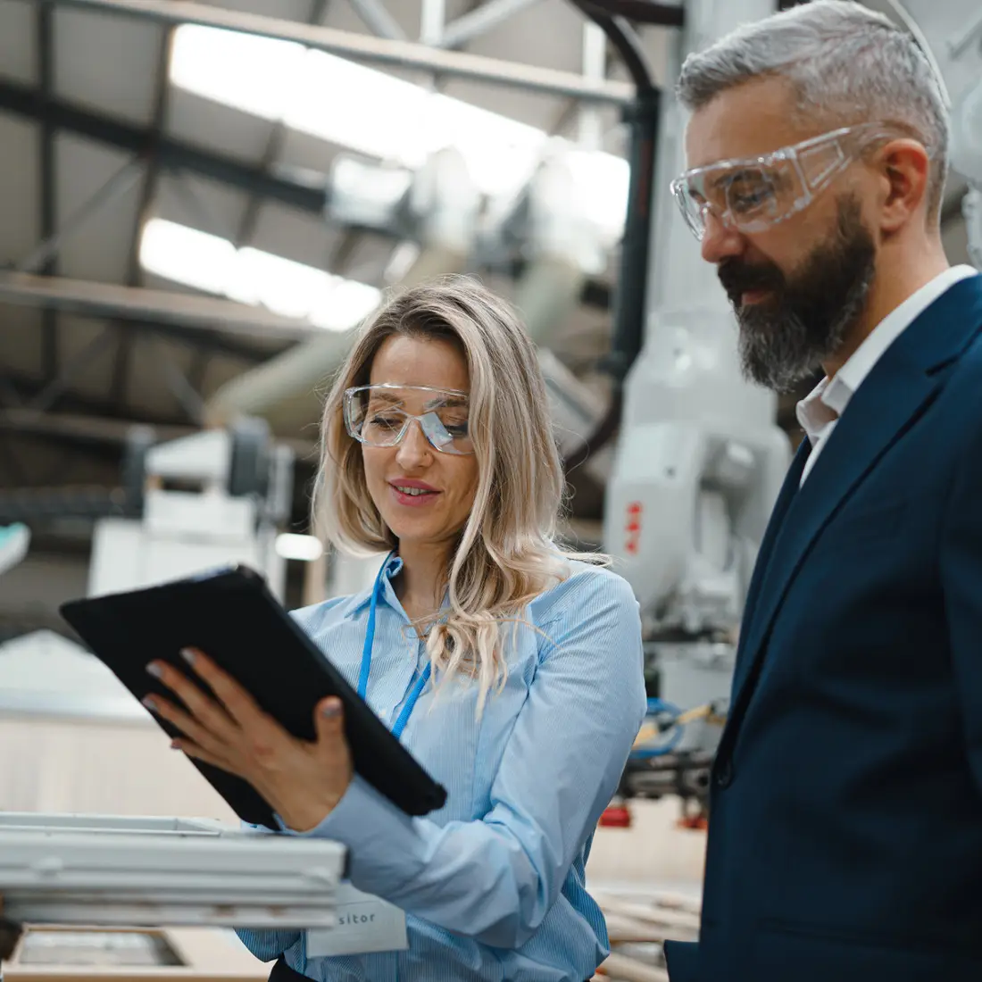 Two people dressed in business attire in a factory wearing safety glasses looking at a tablet