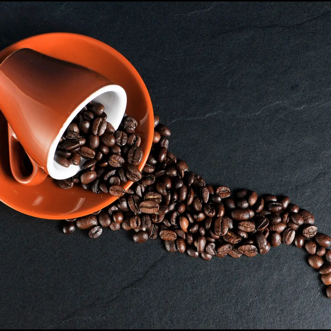 A red coffee cup rests on it's side on a black table with coffee beans spilling out.