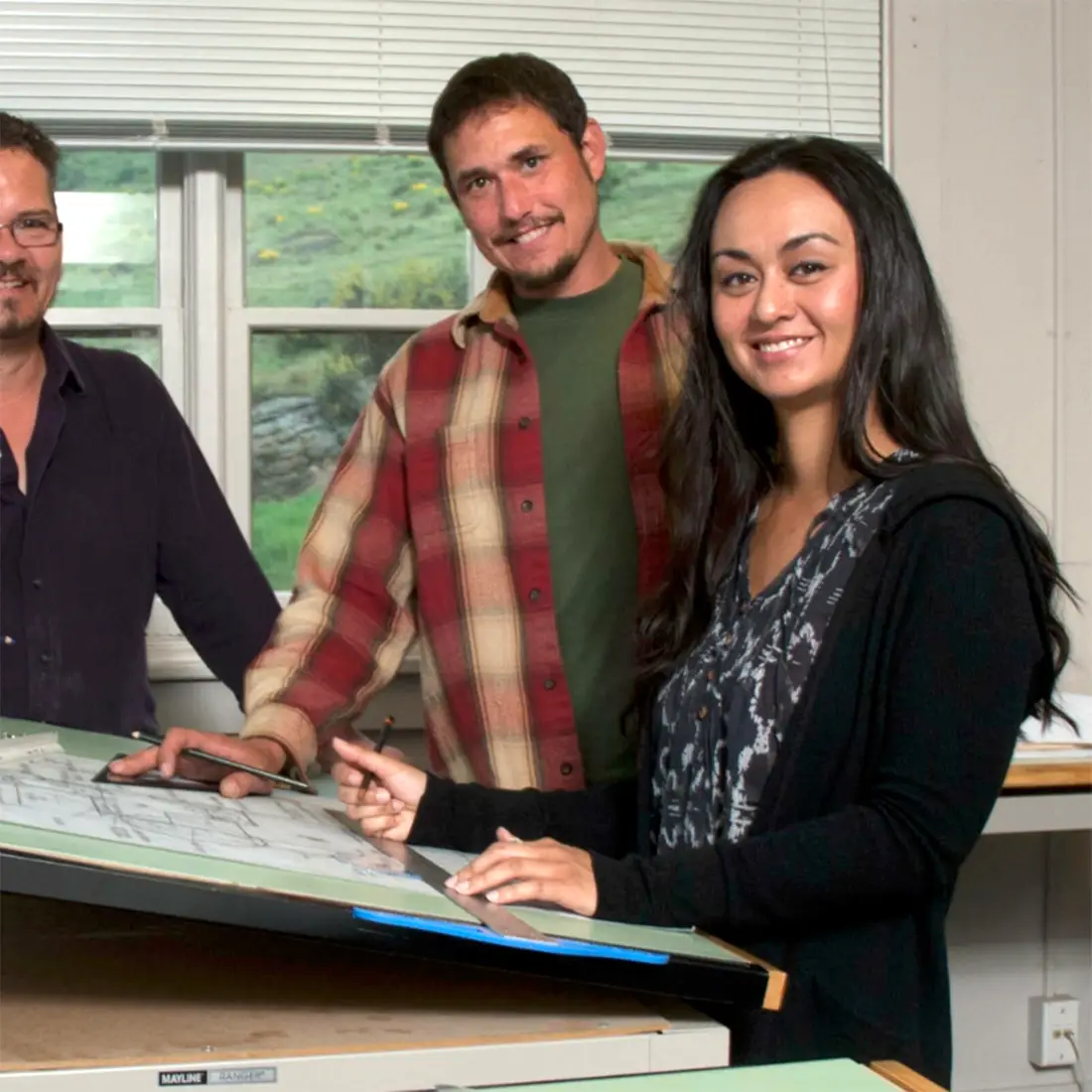 Two entrepreneurs work at a table covered in blue prints alongside a business advisor.