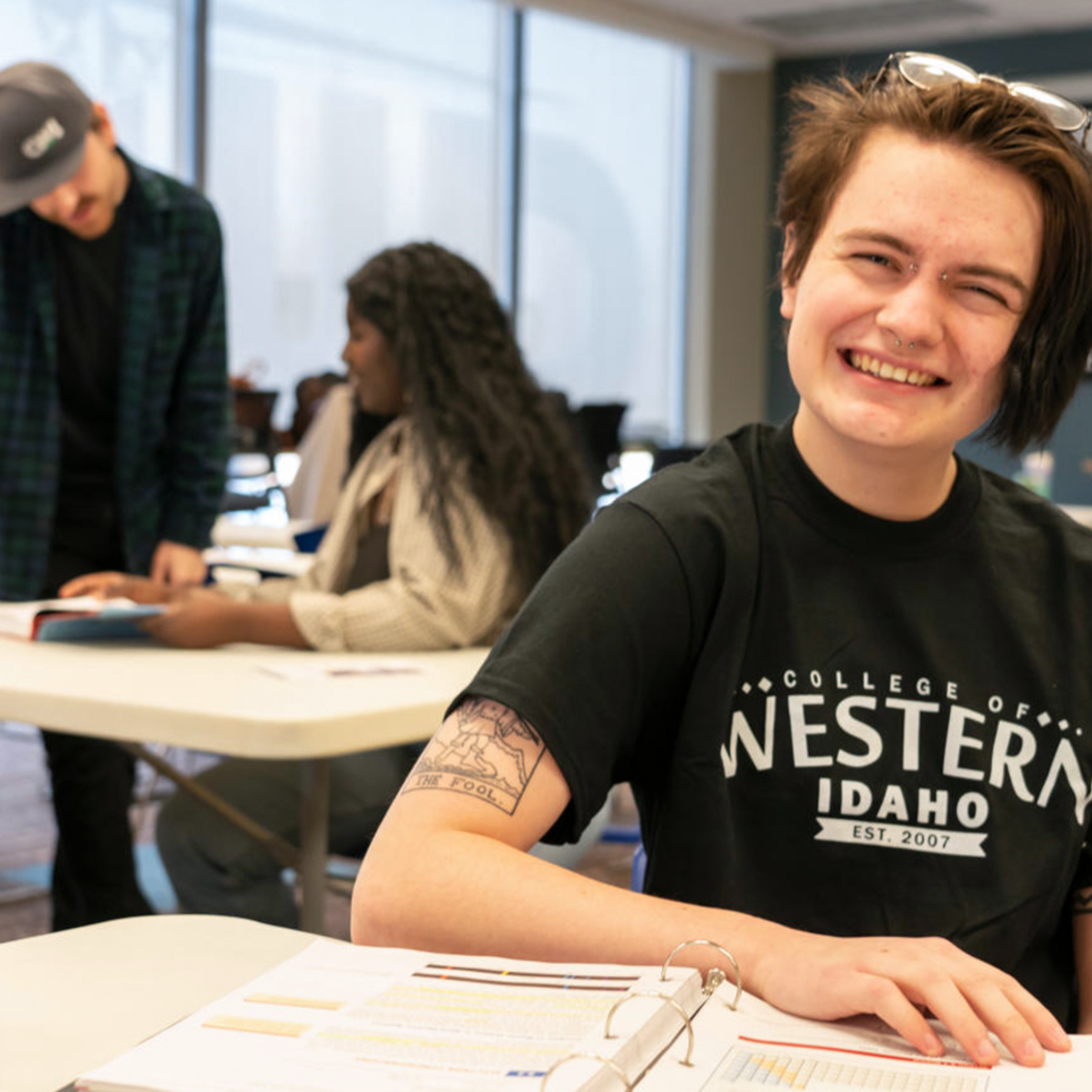 Student in a classroom smiling at the camera. Two students in the background working together.