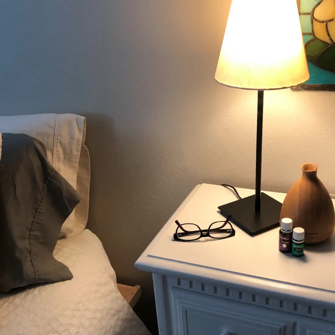 Nightstand next to a bed with glasses, a lamp, and essential oils on it.