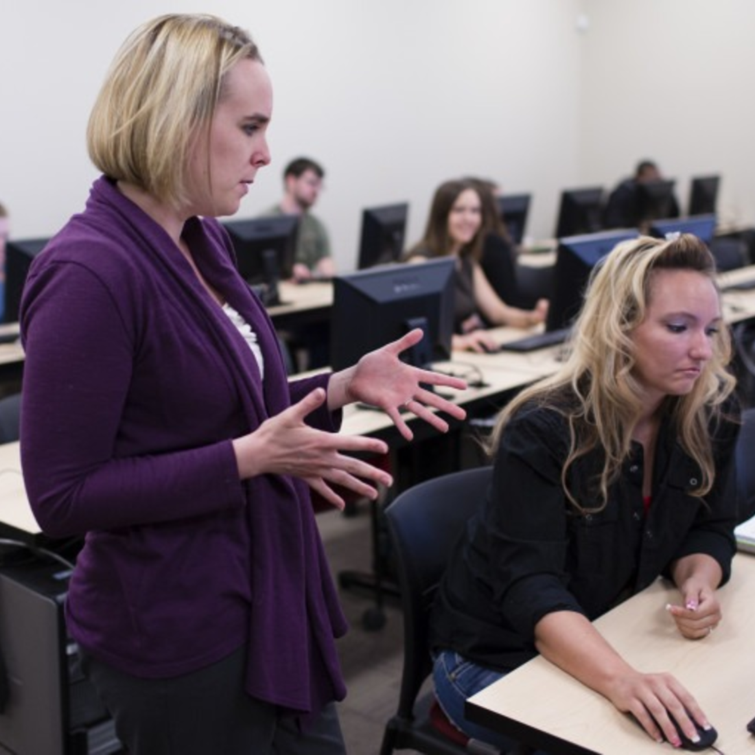 Instructor talking with a student sitting at a computer in a computer lab