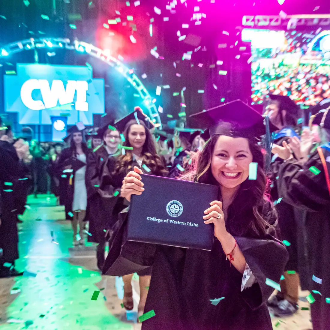 A graduated student wearing cap and gown holds up her new diploma as confetti falls from above.