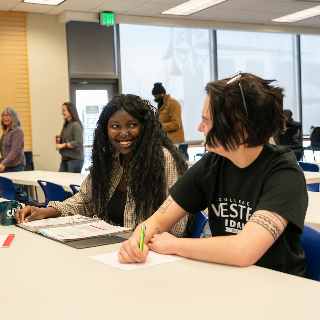 Two students sit at a classroom table smiling and talking while studying, with notebooks and papers in front of them