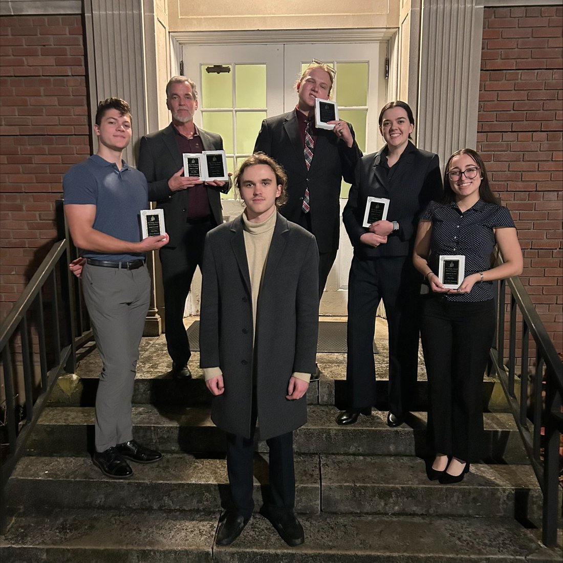 Six students stand on the steps in front of a building with awards