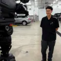 Man stands in an automotive workshop while being filmed, with cars on lifts in the background.