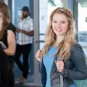 student wearing a backpack smiling at the camera, other students walking into a campus building behind