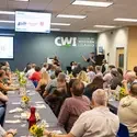 Rows of people sit in front of tables, while watching speaker