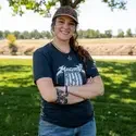 Person stands in front of tree and grass with arms crossed