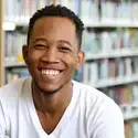 Happy student from Jamaica wearing a white t-shirt sitting in front of shelf of books