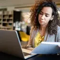 A college student sits in a library in front of a laptop while holding a notebook.