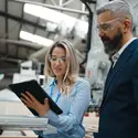 Two people dressed in business attire in a factory wearing safety glasses looking at a tablet