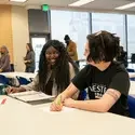 Two students sit at a classroom table smiling and talking while studying, with notebooks and papers in front of them