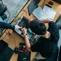 A student bends over a table while looking down into an open computer case. A teacher provides instruction.