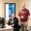 Two workers stand next to a desk posing for a photo. 