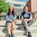two students smiling at the camera sitting outside a campus building on steps