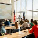 A group of people sit in a bright classroom listening to a speaker during Veterans Day Activities