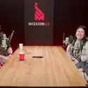 Four people sit at a table in front of microphones and talk while recording a podcast.