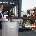 A man at a podium signals to raise a metal construction beam on a work site.