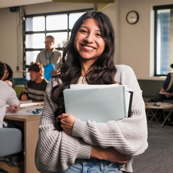 student holding notebooks smiling at the camera inside a classroom with students in the background