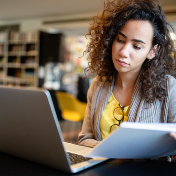 A college student sits in a library in front of a laptop while holding a notebook.