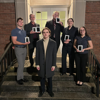 Six students stand on the steps in front of a building with awards