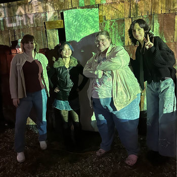 Four people pose together in front of a wooden wall illuminated by colorful, projected light patterns at night.