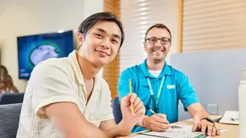 Staff helping a student at a table inside a campus building, both smiling at the camera