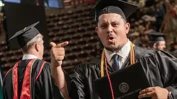 Student in a cap and gown holding and pointing to their diploma