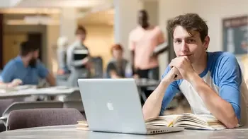 Student sitting at a table in common area on campus with a laptop