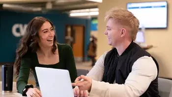 Two students sitting in common area inside a campus building working on a laptop together and smiling at each other