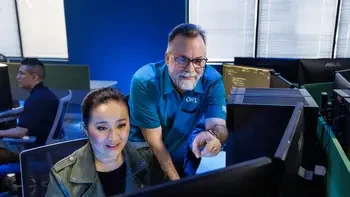 Instructor helping a student in a computer lab at a desktop computer with a student working in the background