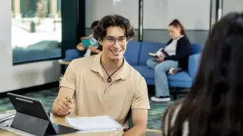 student sitting at a table in common area on campus talking with someone in front of a laptop and notebook
