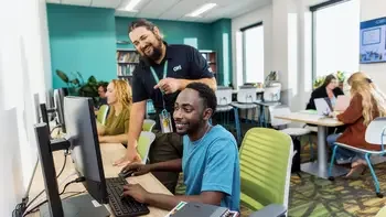 Staff helping a student at a computer. Other students working in the background.