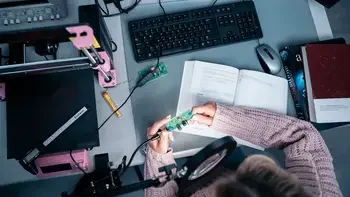 Top down view of a student working at a computer, holding tech items, with books on the desk