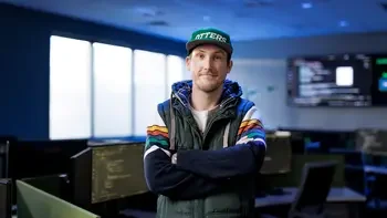 Student standing with arms crossed, wearing an Otters hat, standing in a computer classroom, posing for the camera.